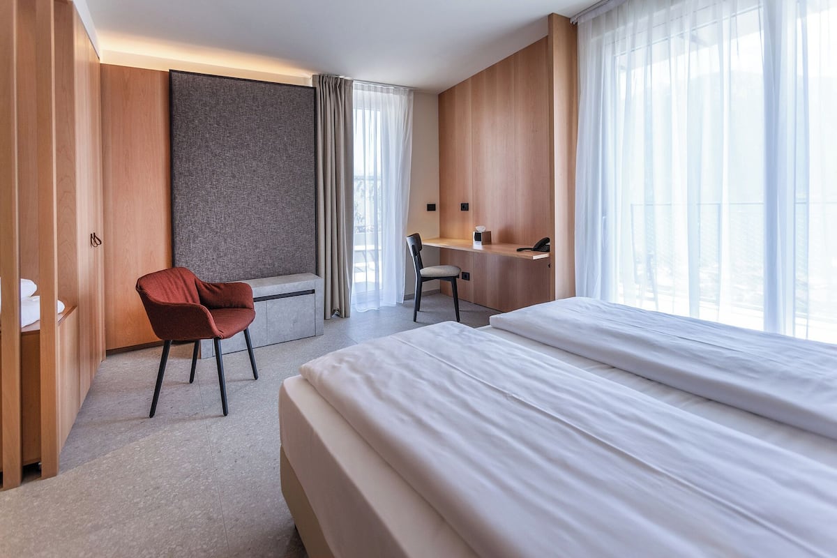 A modern hotel room with a large bed, a desk, a chair, and a wardrobe.
