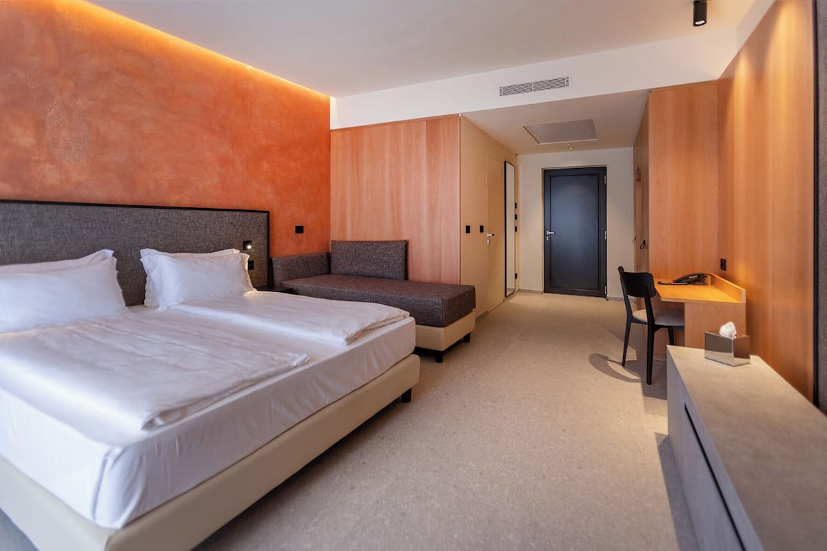 A modern hotel room with a large bed, a sofa, a desk, and a door.