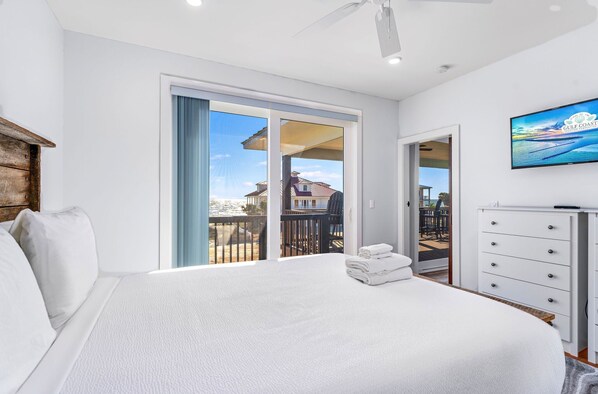 A bedroom with a large bed, a dresser, a TV, and a balcony with a view of houses. - Dragonfly Shores - Beach View, Pet Friendly, Beach Gear Credit Included! (St George Island)
