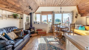 A cozy living room with a fireplace, a dining area, and a large window offering a view. - 3 Bed in East Prawle (oc-t30117) (East Prawle)