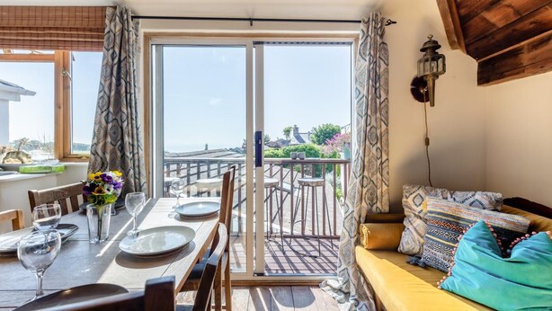 A dining area with a table set for two, a sofa with patterned cushions, and a balcony with a railing. - 3 Bed in East Prawle (oc-t30117) (East Prawle)