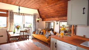 A cozy room with a wooden ceiling, a sofa with cushions, a dining table with chairs, and a kitchen area with a dishwasher. - 3 Bed in East Prawle (oc-t30117) (East Prawle)