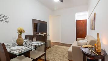 A dining area with a glass table, chairs, and a vase with flowers. There is a television mounted on the wall and a wooden door in the background.