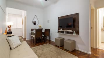 A living room with a sofa, a TV mounted on the wall, a dining table with chairs, and a small coffee table.