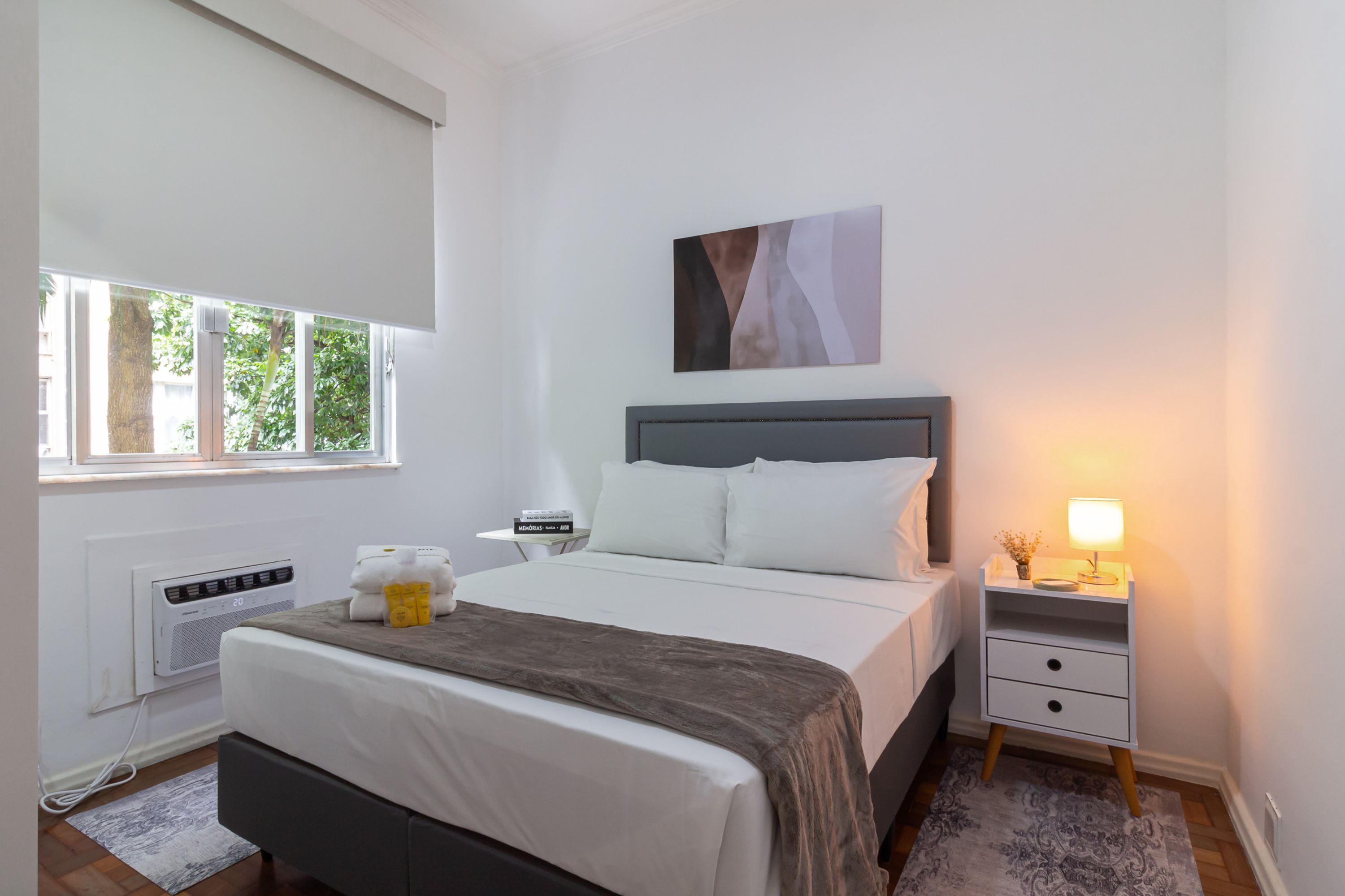 Een slaapkamer met een bed, nachtkastje en een raam met uitzicht op bomen.