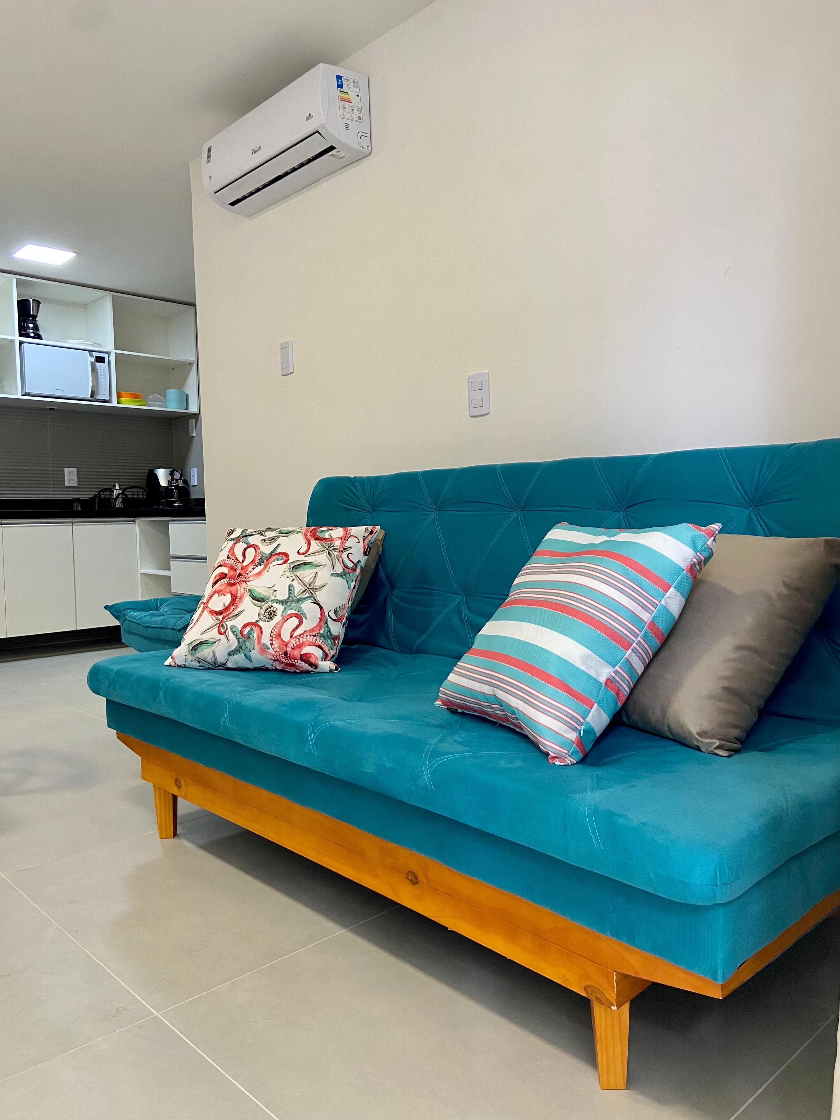 Un sofá color turquesa con patas de madera, dos cojines estampados y un aire acondicionado blanco montado en la pared.