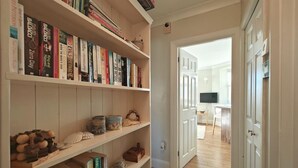Un pasillo con una estantería llena de libros, piso de madera y una puerta que da a otra habitación con televisor y una pequeña mesa.