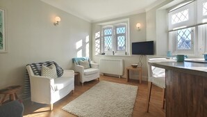 Una sala de estar moderna con sofá, sillón y televisor. Se ven ventanas con persianas y un radiador.