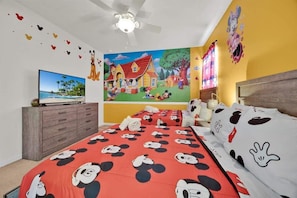 A bedroom with a Mickey Mouse themed bedspread, a large mural on the wall, and a ceiling fan.