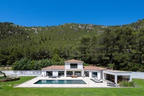 A modern two-story house with a swimming pool, surrounded by a well-maintained lawn and dense forest.