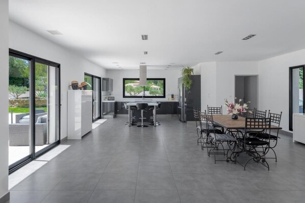 A modern kitchen with a large island, a dining area with a wooden table and metal chairs, and large windows offering a view of the outdoors.