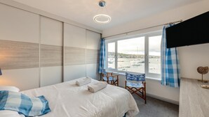 A bedroom with a bed, a chair, a TV, and a window with a view of boats and a beach.