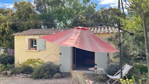 Een klein huis met een terras, voorzien van twee stoelen en een tafel onder een rode parasol.