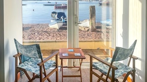 A balcony with two chairs and a table, overlooking a calm harbor with boats and a sandy beach.
