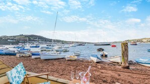 A beachside view with boats docked in the water, a sandy shore, and a table with a glass and a bottle.