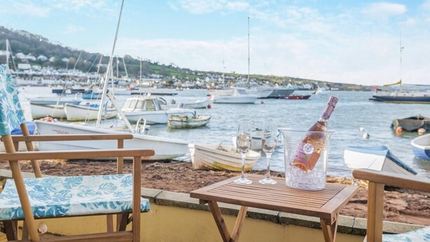 A beachside view with wooden chairs and a table set with a bottle and glasses, overlooking a marina with boats.