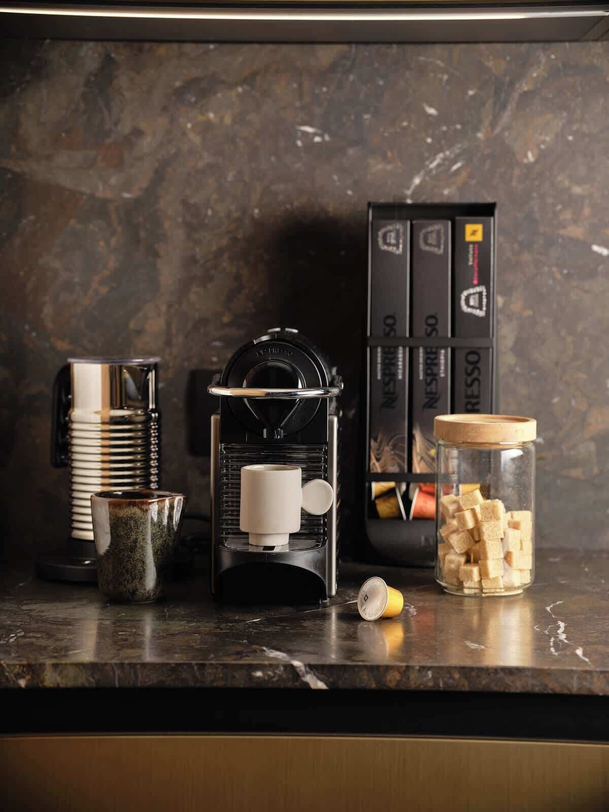 A coffee machine, a stack of cups, a jar of sugar cubes, and a coffee pod box on a kitchen counter.