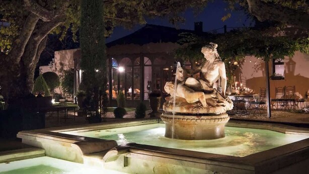 Une fontaine ornée d’une statue, entourée d’un bassin et d’arbres, avec un bâtiment en arrière-plan.