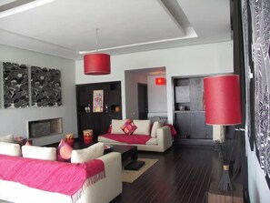 A modern living room with a sofa, coffee table, and red lamps. There is a fireplace and artwork on the wall. - Villa Sylana 2 Pools Tennis Court Hammam (Marrakesh)
