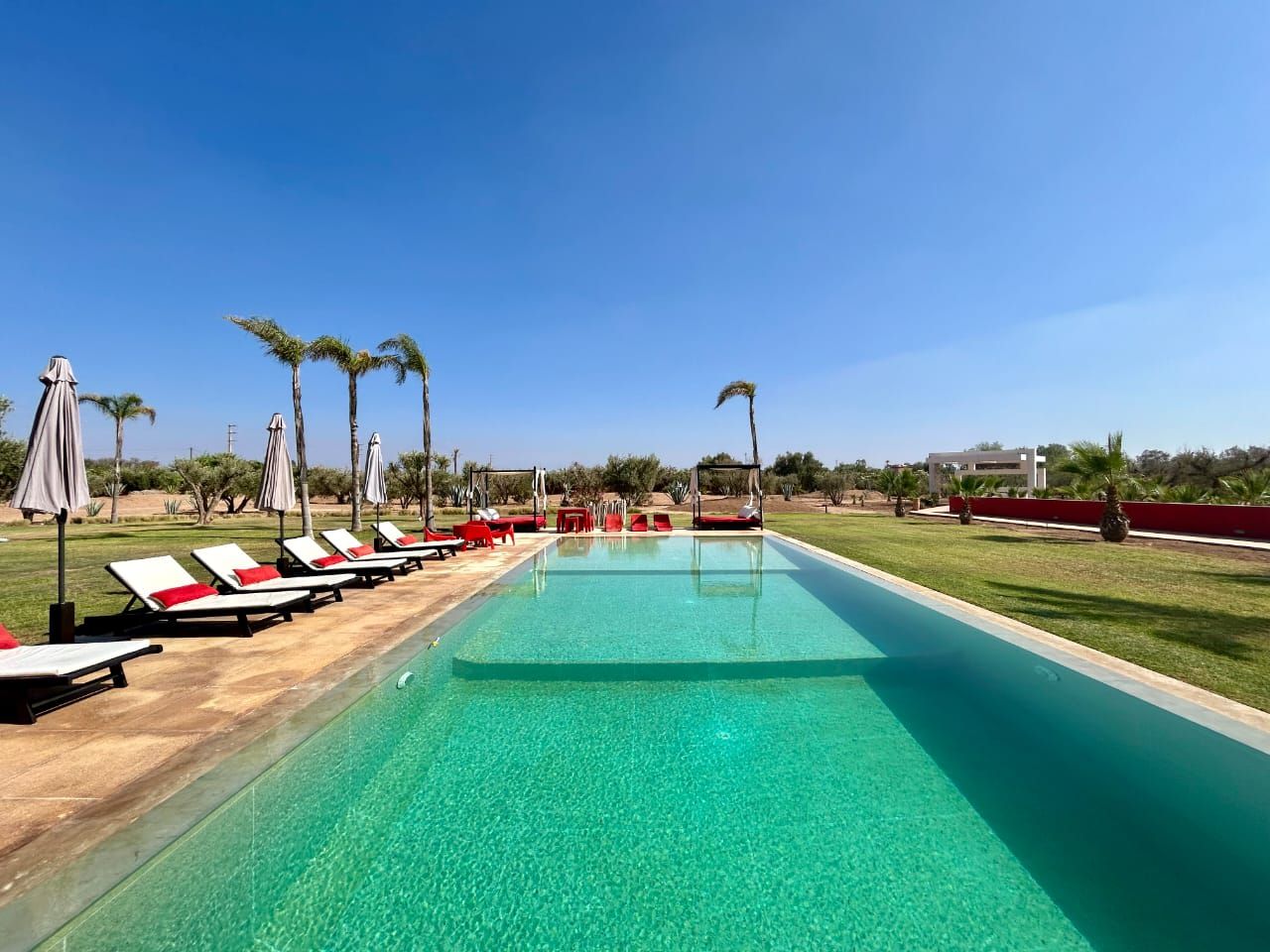 A swimming pool with lounge chairs and umbrellas, surrounded by greenery and a clear sky.