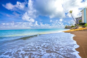 A beach with clear blue water, waves, and a sandy shore. There is a building with balconies and palm trees in the background.