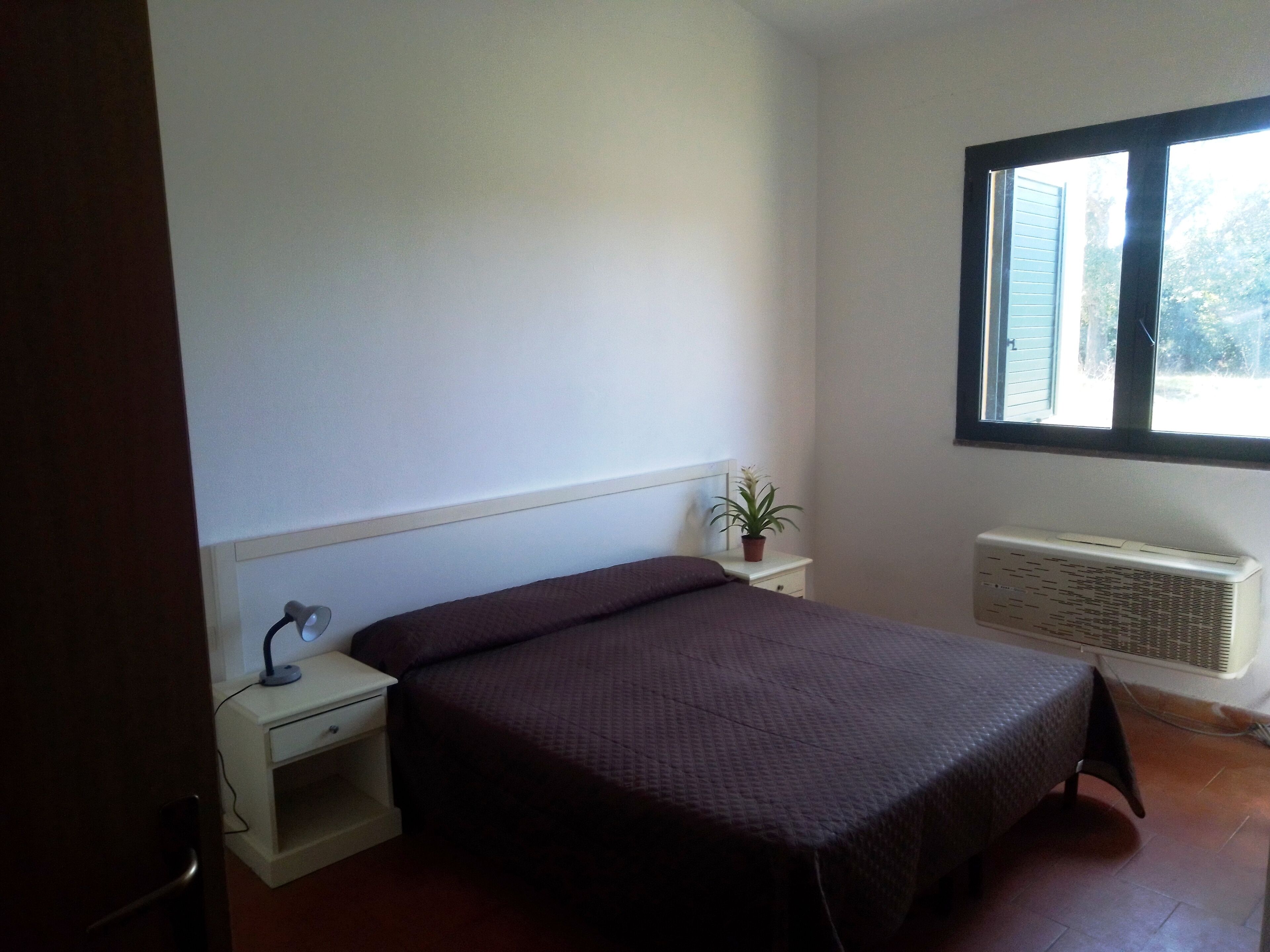 Een slaapkamer met een bed, nachtkastje, lamp, potplant, raam en een airconditioner.