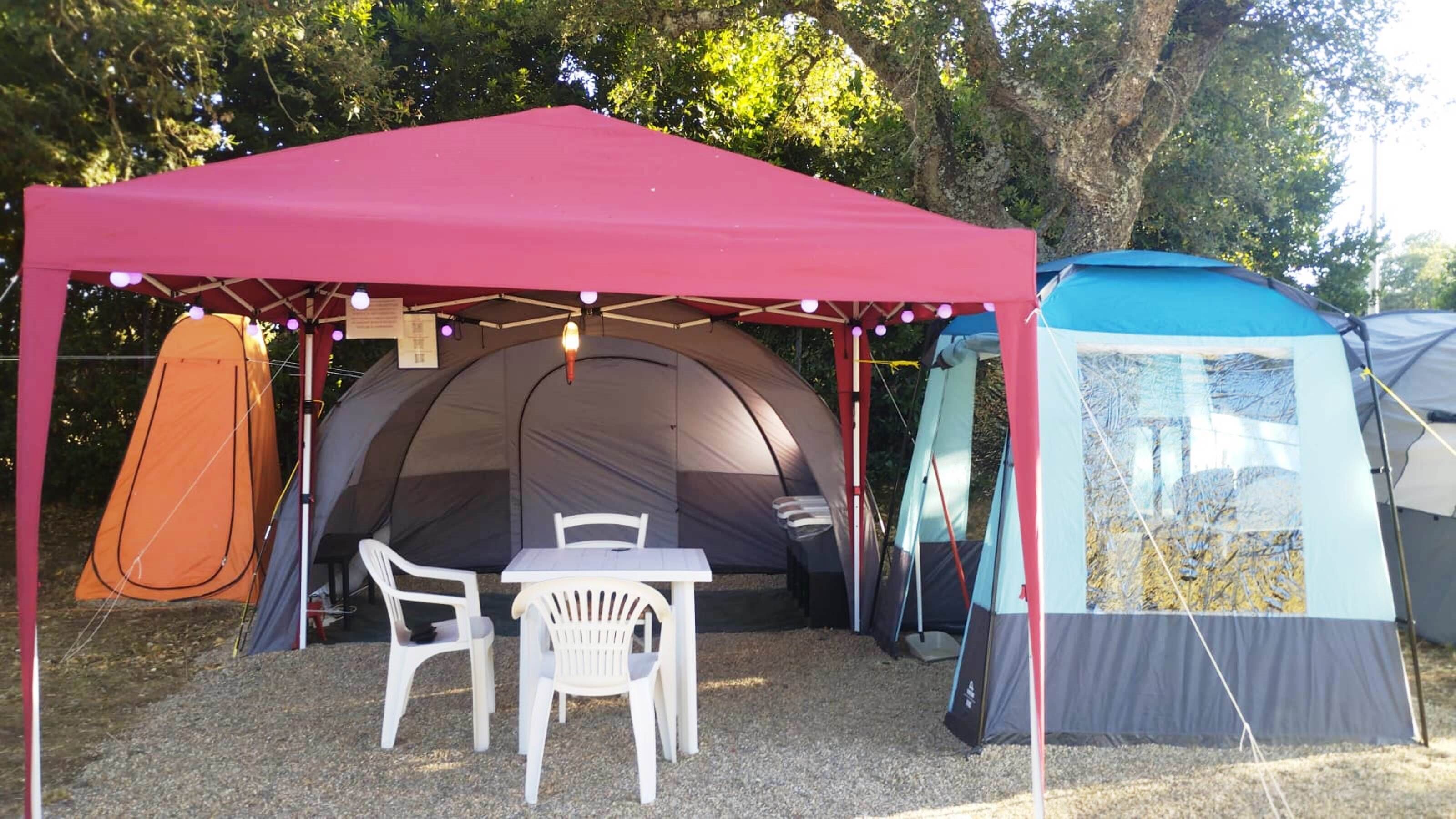 Instalación de campamento con una tienda grande, una tienda más pequeña y dos sillas de plástico blancas bajo un toldo rojo.