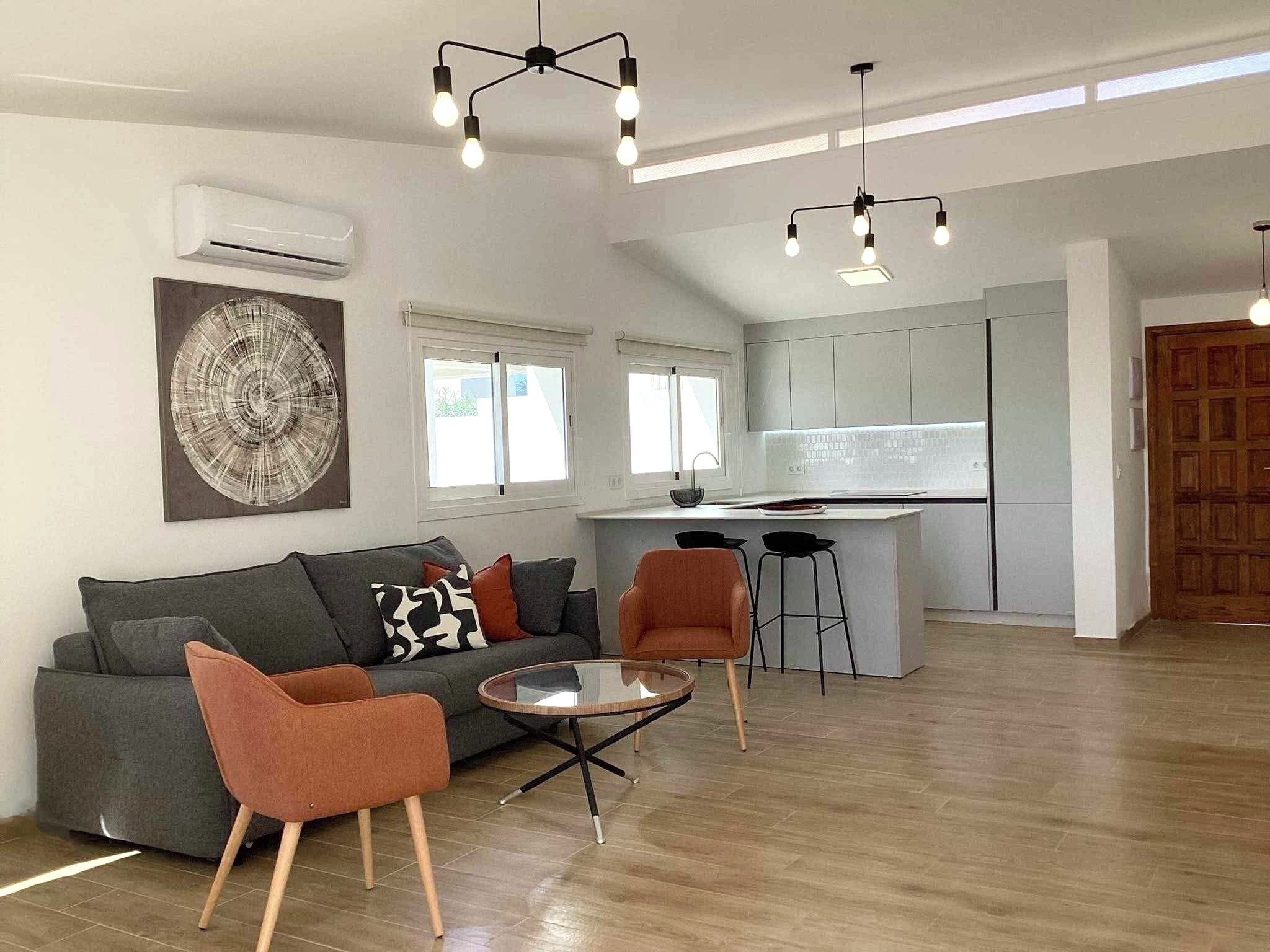 A modern living room with a grey sofa, orange chairs, a glass coffee table, and a kitchen in the background.