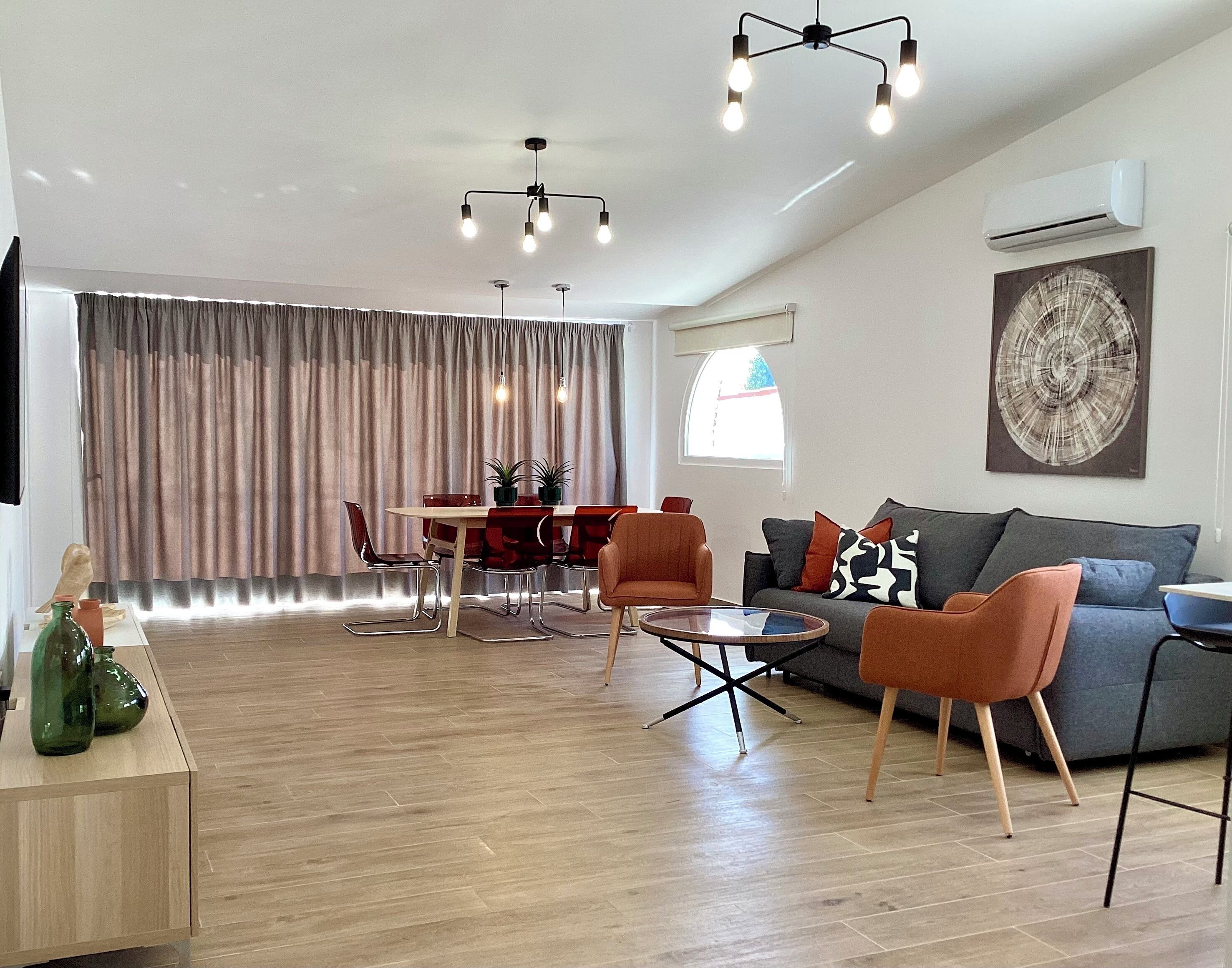 A modern living room with a dining area, featuring a wooden floor, a grey sofa, red chairs, and a dining table with chairs.
