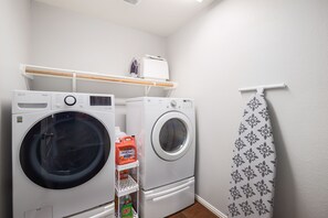 Uma lavanderia com duas máquinas de lavar brancas de carregamento frontal, uma prateleira com produtos de lavagem e um chuveiro com cortina estampada.
