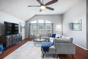 A spacious living room with a ceiling fan, a flat-screen TV, a gray sofa, a coffee table, and a large window with curtains. - Executive 4BR Round Rock Stay | Near Dell (Round Rock)