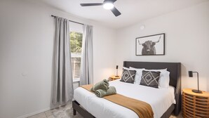 Een slaapkamer met een bed, een plafondventilator, een raam met gordijnen, een aan de muur bevestigd schilderij en een bedlampje.