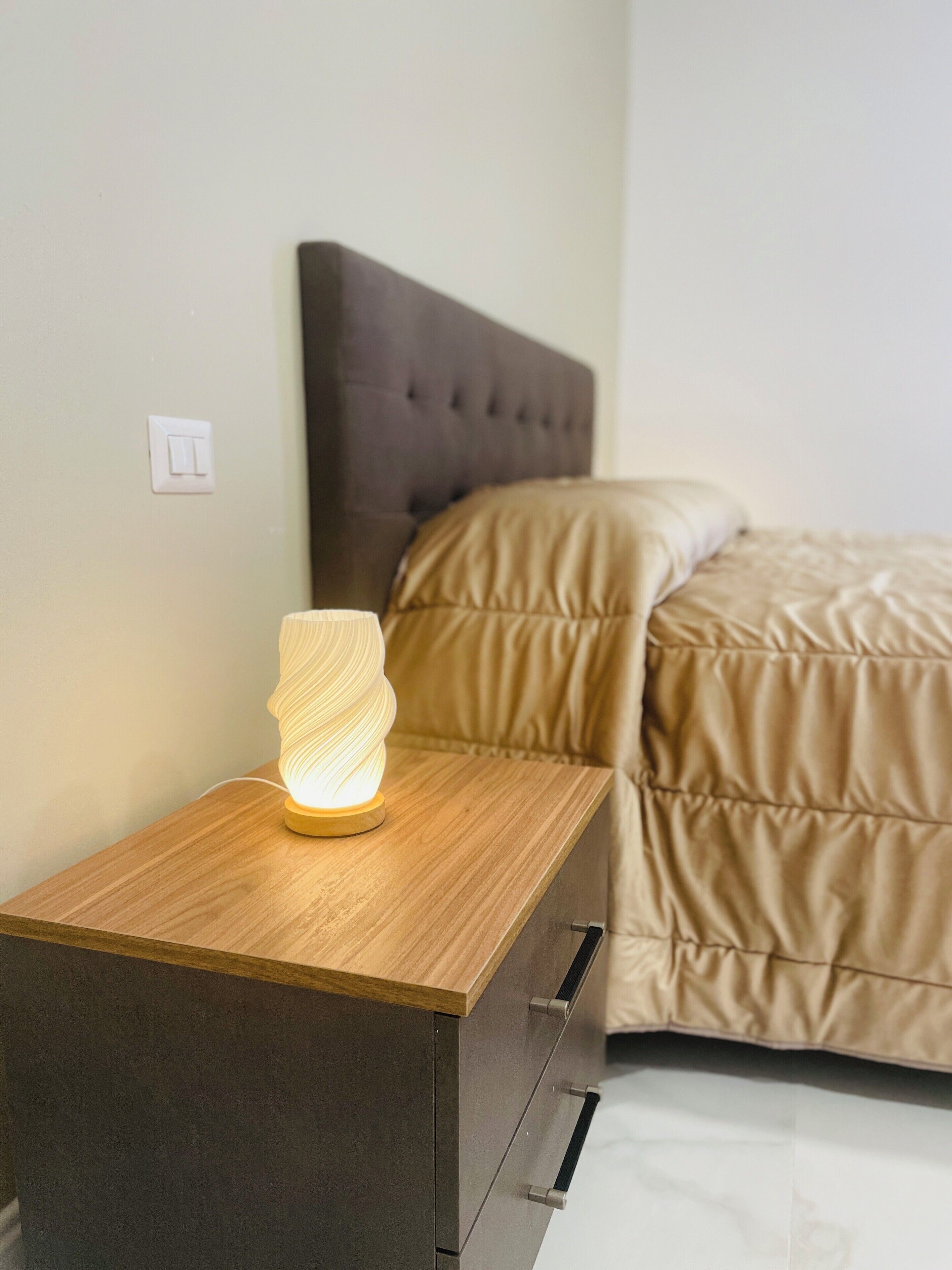 Una mesita de noche con lámpara, una cama con cabecero e interruptor de pared.