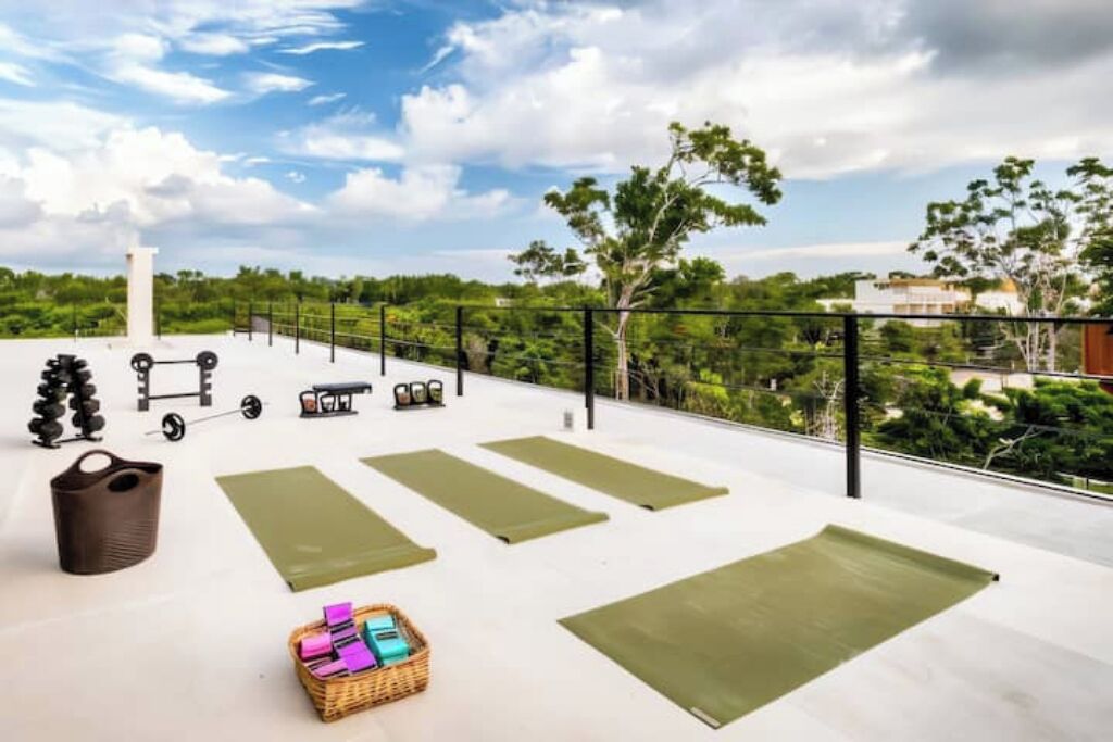 A rooftop gym with exercise equipment and mats, surrounded by greenery and a clear sky.