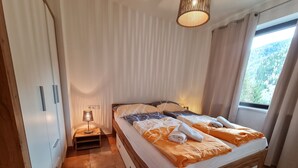 A bedroom with a wooden bed, a bedside table with a lamp, a window with curtains, and a view of trees outside. - Apartment With Mountain & Slope View (Bad Kleinkirchheim)