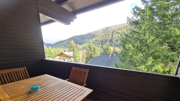 A wooden table and chairs on a balcony with a view of a forested valley and a house in the distance. - Apartment With Mountain & Slope View (Bad Kleinkirchheim)