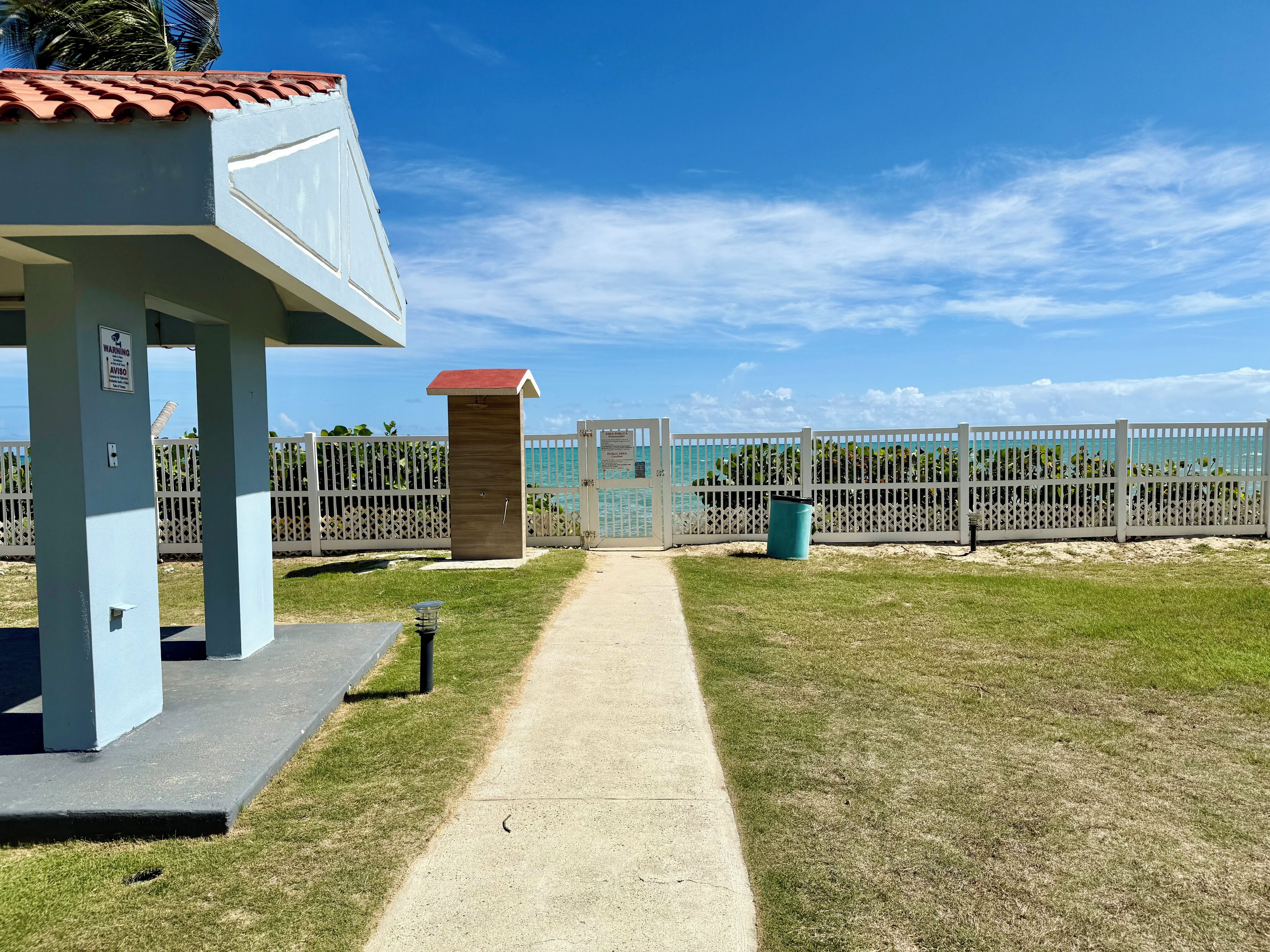 A pathway leading to a building with a red roof, a fenced area with a gate, and a clear sky.