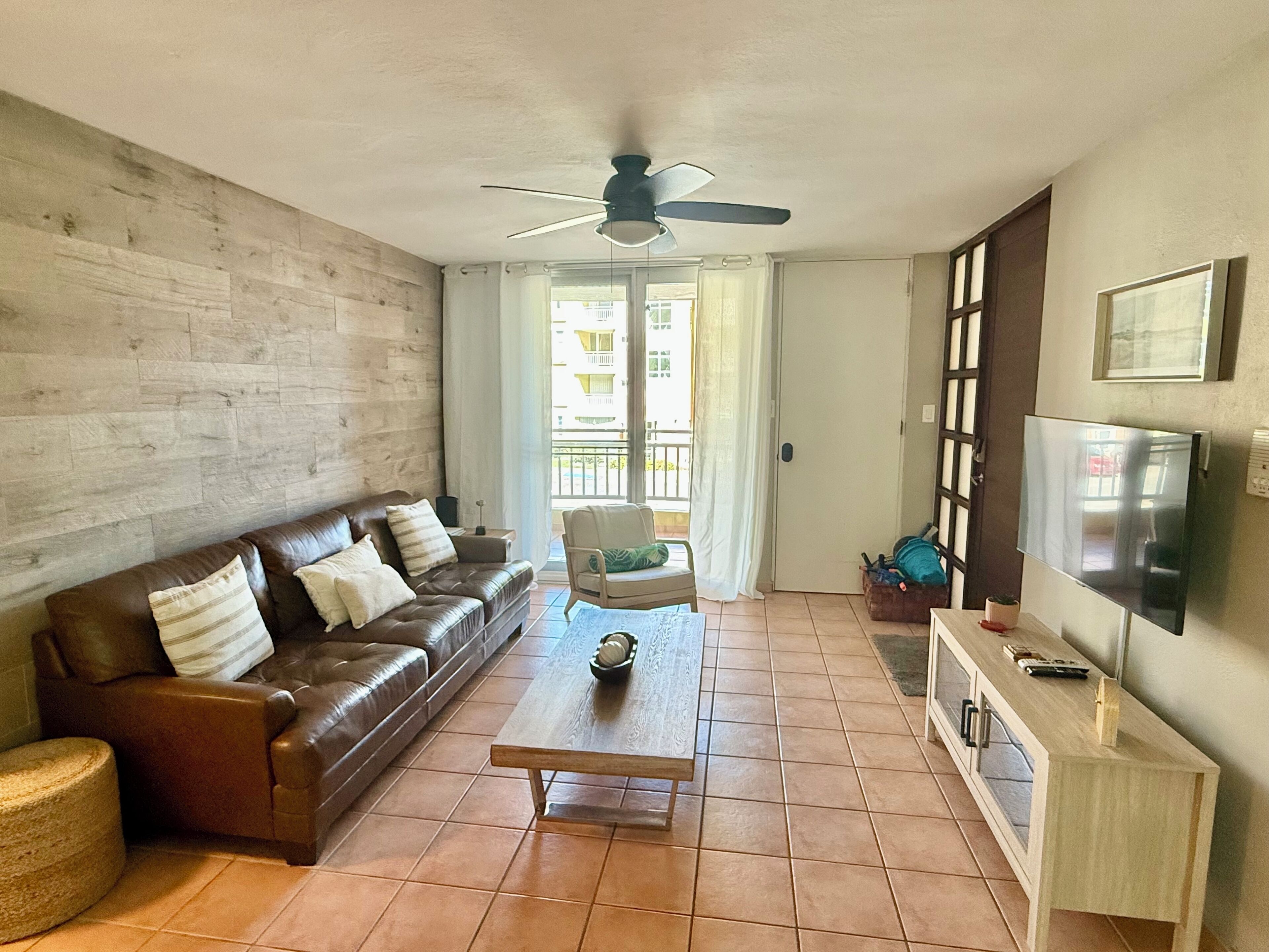Un salon avec un canapé en cuir marron, une table basse en bois, un ventilateur de plafond, un téléviseur posé sur un meuble en bois et une porte donnant sur un balcon.