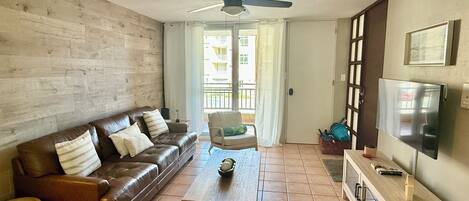 Un salon avec un canapé en cuir marron, une table basse en bois, un ventilateur de plafond, un téléviseur posé sur un meuble en bois et une porte donnant sur un balcon.