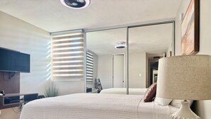 Un dormitorio moderno con una cama grande, una mesita de noche con lámpara, un televisor montado en la pared y un espejo grande que refleja la habitación.