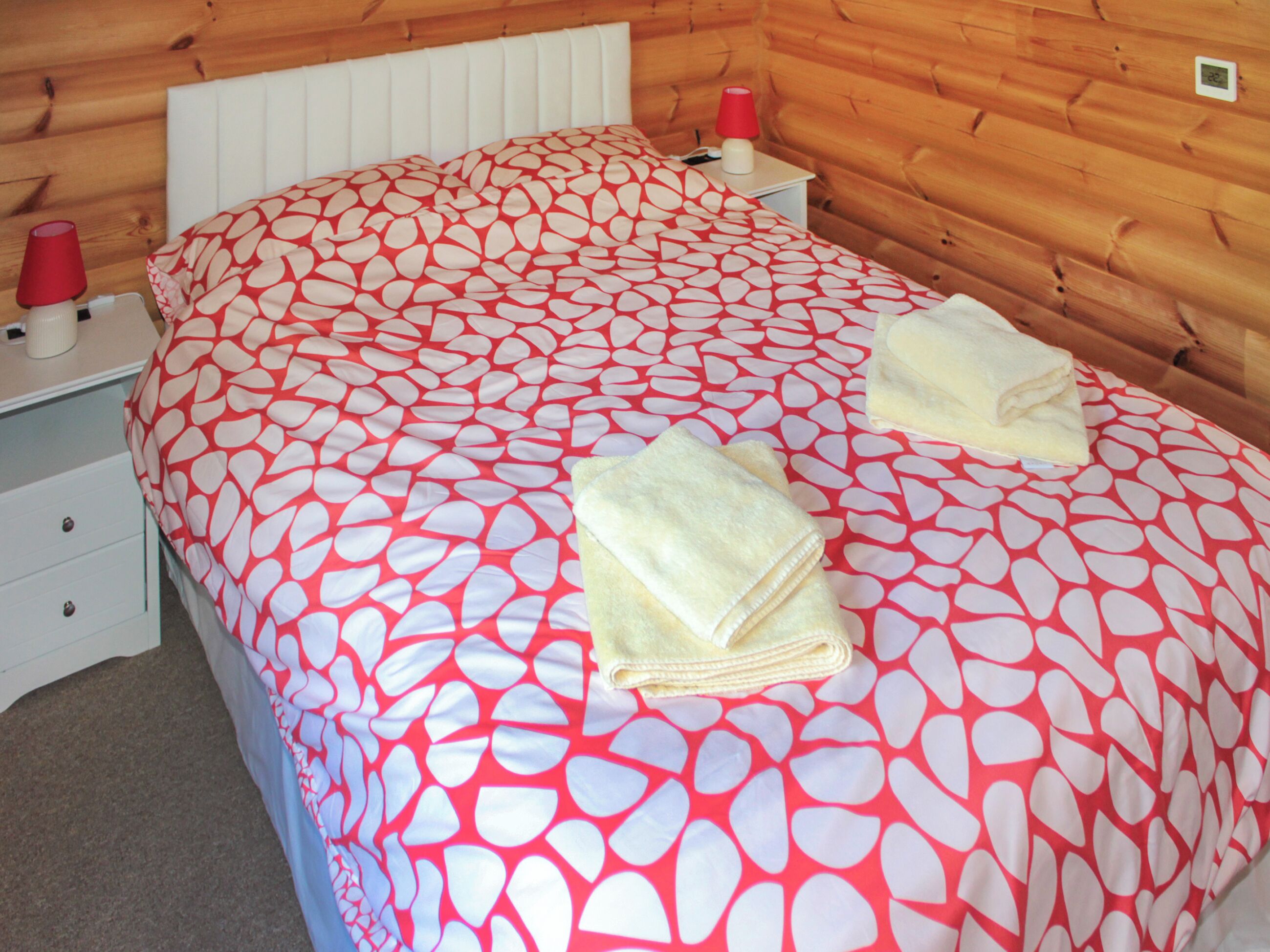 Una habitación con una cama cubierta por una colcha roja y blanca, dos toallas blancas dobladas con cuidado sobre la cama, una mesita de noche blanca con una lámpara roja y un mueble blanco con cajones.