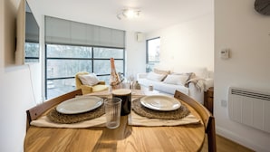 A dining area with a wooden table set for two, a large window with blinds, a white sofa, and a clock on the wall. - Modern Flat, Oxford city centre (Oxford)