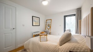 A bedroom with a large bed, a bedside table with a lamp, a window with curtains, and a door. - Modern Flat, Oxford city centre (Oxford)
