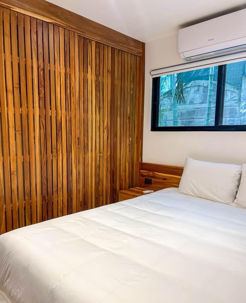 Een slaapkamer met een wand van houten latten, een bed met wit beddengoed en een raam met uitzicht op groen.