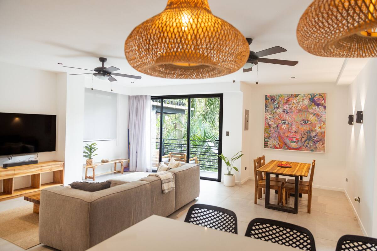 Una sala de estar moderna con sofá, mesa de comedor, sillas, televisor y un ventanal con vistas a la vegetación.