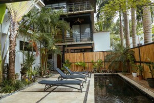A modern outdoor pool area with lounge chairs, a wooden fence, and tropical plants.