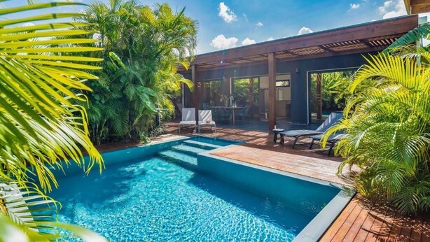 Una piscina con muelle de madera y sillas reclinables, rodeada de abundante vegetación.