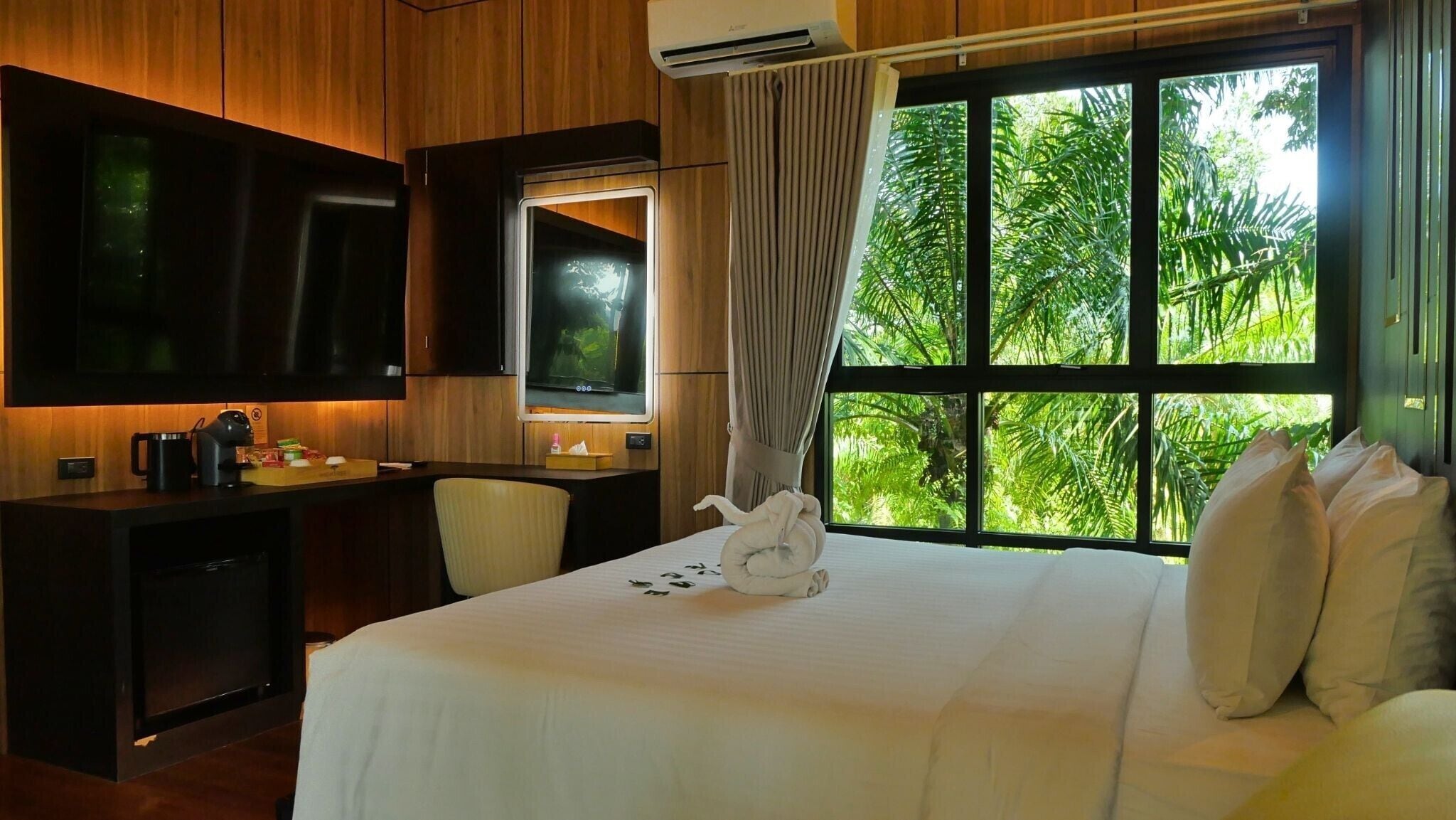 A hotel room with a bed, a desk, a chair, a TV, and a window with a view of greenery.