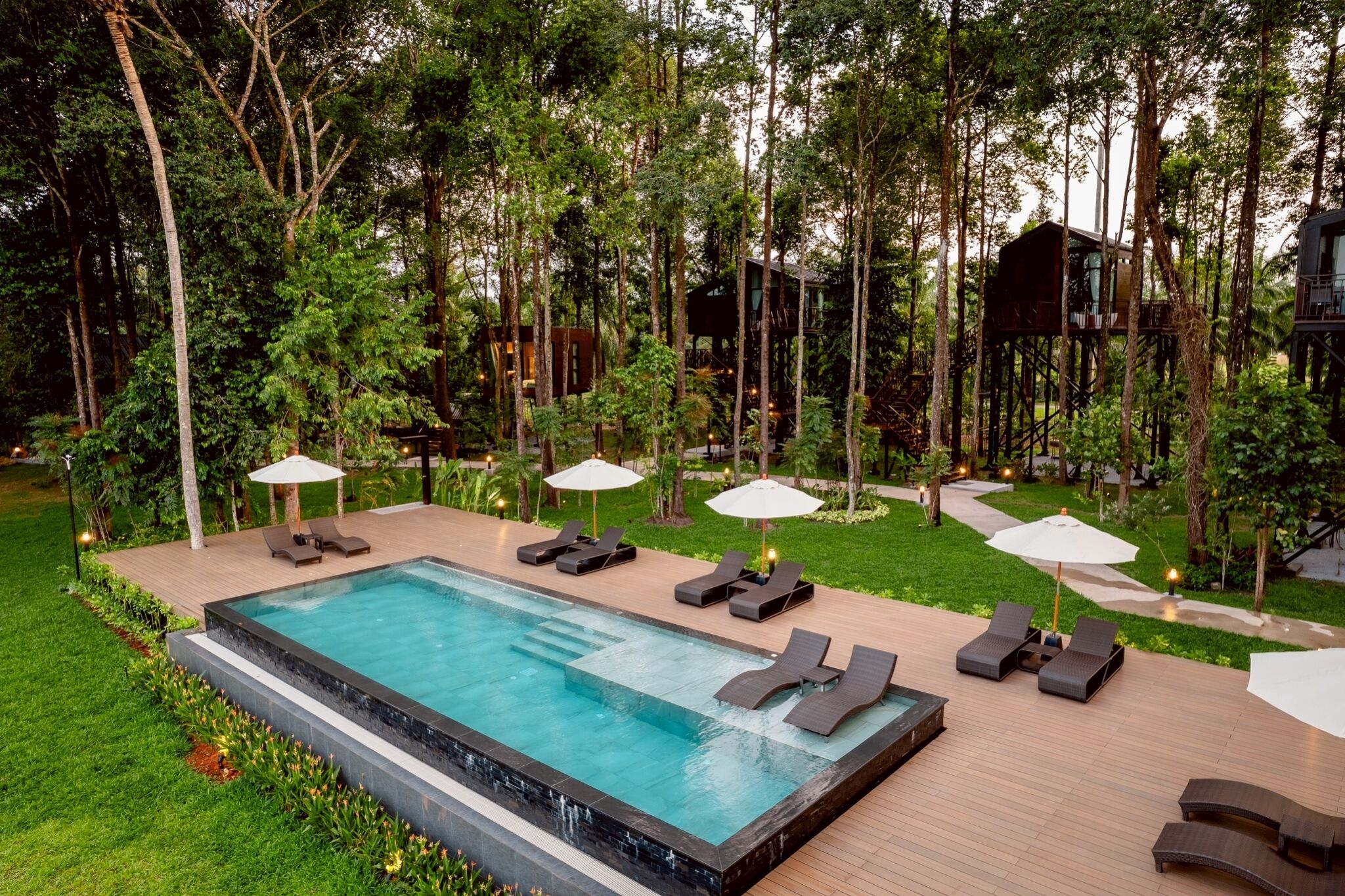 Piscina com espreguiçadeiras e guarda-sóis, rodeada por árvores e um deck de madeira.