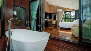A modern bathroom with a freestanding bathtub, wooden flooring, and large windows offering a view of the bedroom.